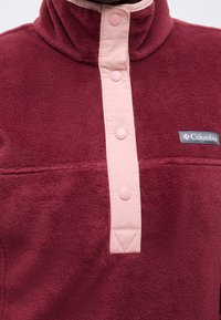 Donkerrode fleece trui met een roze knoopsluiting en een grijs Columbia logo op de borst.