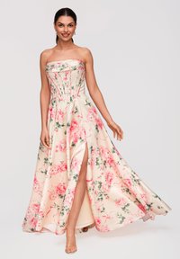 Femme souriante portant une robe fleurie crème sans bretelles avec des fleurs roses et une fente haute, posant devant un fond clair.