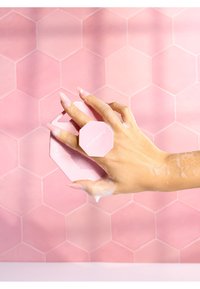 Hånd med lange negle, der holder en lyserød sekskantet skrubbesvamp mærket "nashworks," dækket af sæbebobler mod en baggrund af lyserøde sekskantede fliser.