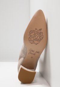 Zapato de tacón alto de ante beige exhibido de lado. Presenta una suela texturizada con el logo "Apple of Eden" y la frase "Sands from Paradise", talla 37.