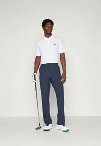 Άνδρας που φοράει λευκό πόλο, ναυτικά παντελόνια και γάντι γκολφ κρατάει ένα κλαμπ γκολφ, στέκεται μπροστά από απλό ανοιχτό φόντο.