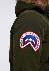 Green winter jacket with a fur-lined hood. Features a round embroidered patch in red, white, and blue on the left sleeve.