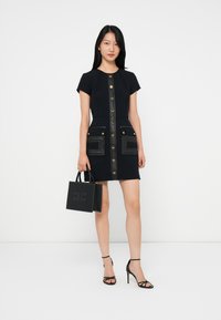 Mujer de pie con un vestido negro de manga corta y botones frontales, sosteniendo un bolso negro con tachuelas, llevando sandalias de tacón alto negras, sobre un fondo blanco.