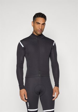 Mann in schwarzer langärmliger Radjacke und Hose mit reflektierenden Streifen an den Schultern und Oberschenkeln, stehend vor einem einfarbigen Hintergrund.