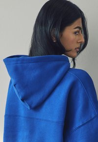 Hoodie i klarblått, tillverkad av mjukt material. Har ribbad kant och kontrasterande sömmar, med en avslappnad passform och en stor huva.