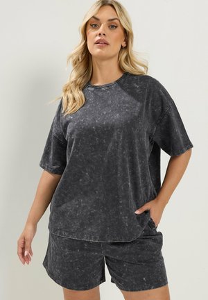 Femme blonde portant un t-shirt noir oversize effet délavé et un short assorti, debout avec une main dans la poche sur un fond clair.