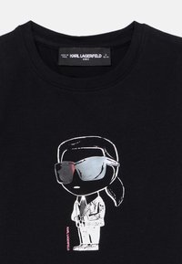 Μαύρο βαμβακερό T-shirt με καρτούν σχέδιο μιας φιγούρας με γυαλιά ηλίου και κοστούμι, με λευκό περίγραμμα και ροζ κείμενο "KARL LAGERFELD."