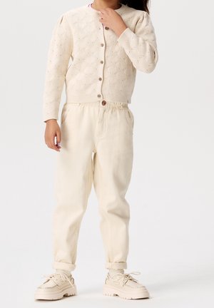 Cardigan en maille ivoire avec motif en vagues, fermeture par boutons, associé à un pantalon beige à taille élastique et des chaussures beige clair. Texture douce.