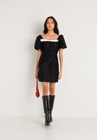 Černé šaty s vypufovanými rukávy a kontrastním bílým výstřihem, strukturovaná látka, doplněné černými kozačkami po kolena a malou červenou kabelkou.