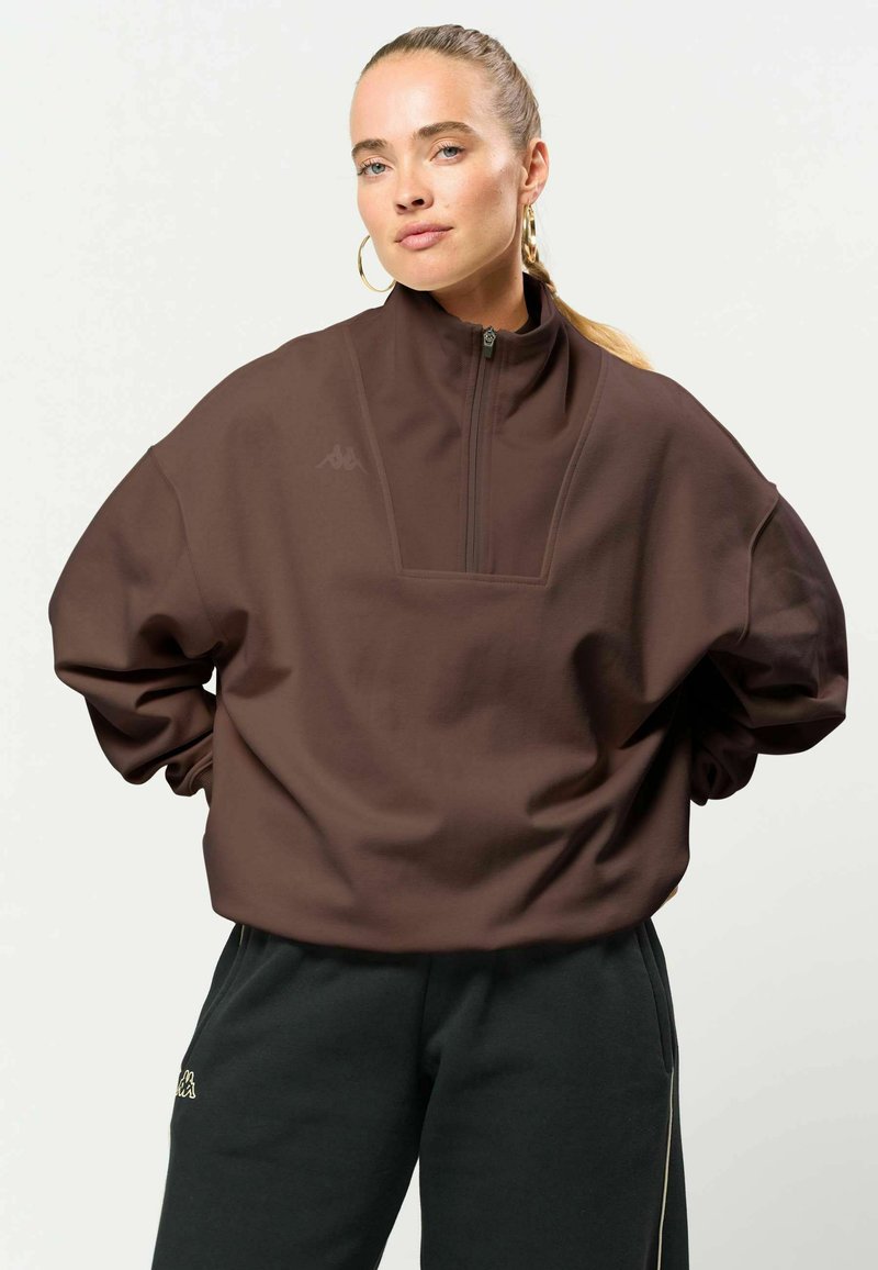 Femme à la peau claire et aux cheveux blonds attachés, portant de grandes boucles d'oreilles en anneau, un pull marron zippé et un pantalon de survêtement noir, debout les mains sur les hanches.