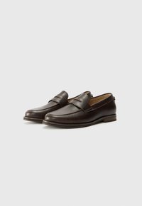 Mocasines de cuero marrón con una textura suave, que presentan un detalle de correa y una punta redondeada, asentados sobre un tacón bajo de madera.