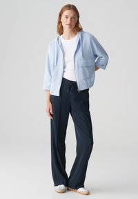 Giacchetto azzurro chiuso con zip e cappuccio, con tasca frontale; abbinato a pantaloni navy a gamba larga e scarpe da ginnastica bianche. Tessuto liscio, vestibilità comoda.