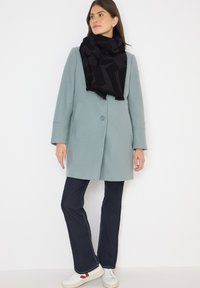 Femme portant un manteau bleu clair, une écharpe noire à motifs, un jean bleu foncé et des baskets blanches, se tenant contre un fond blanc.