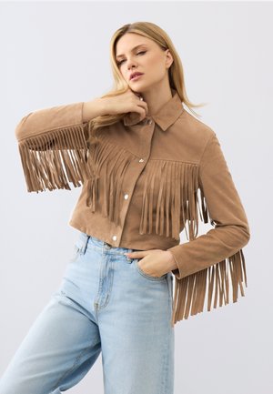 Donna che indossa una giacca di camoscio beige con frange e bottoni d'argento e jeans a vita alta color azzurro chiaro, posa con una mano vicino al viso.