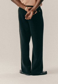 Άτομο που στέκεται με τα χέρια σταυρωμένα πίσω από την πλάτη, φορώντας μαύρο παντελόνι, καφέ μπλούζα, μαύρα παπούτσια και χρυσό ρολόι στον καρπό.