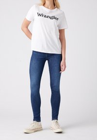 Camiseta de algodón blanca con el logo "Wrangler" en negro, combinada con jeans ajustados azules y zapatillas blancas. Diseño simple, ajuste casual.