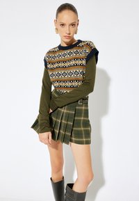 Sweter wełniany z wzorzystym dekoltem w ziemistych kolorach, zestawiony z zieloną spódnicą w kratę z plisami. Modelka nosi czarne kozaki do kolan.