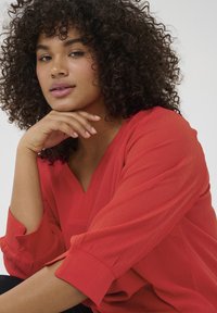 Blouse rouge texturée avec un décolleté en V et des manches trois-quarts. Le modèle a des cheveux bouclés et repose son menton sur sa main.