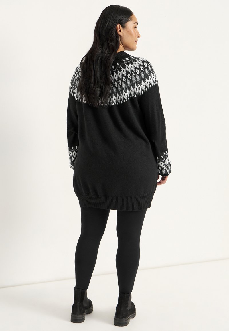 Sort sweater med et strikket mønster i hvidt ved skulderen og ærmerne, med en løs pasform og ribbet kant, parret med sorte leggings og ankelstøvler.