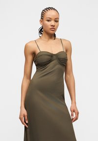 Sieviete ar pītiem matiem, valkājot olīvu zaļas krāsas piedurkņu kleitu, stāv pret vienkāršu gaišu fonu.