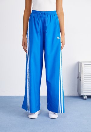 Personne portant un pantalon de survêtement Adidas bleu vif avec des bandes blanches sur les côtés et des baskets blanches, debout sur un sol bleu.
