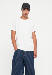 Tricou alb cu mânecă scurtă și un mic logo negru pe piept, asortat cu pantaloni largi cu dungi albastre închise.