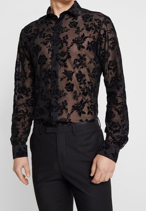 Mężczyzna w czarnej przezroczystej koszuli z kwiatowym wzorem na długim rękawie, włożonej w czarne eleganckie spodnie z paskiem.