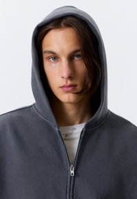 Grå hettegenser med glidelås, stor hette, myk tekstur og sølvfarget glidelås. Brukt over en hvit skjorte, med en synlig sikkerhetsnål detalj.