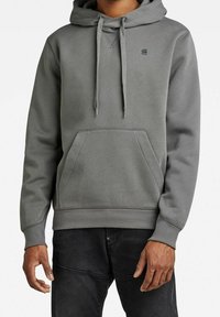 Sudadera gris de tela suave, con un bolsillo tipo canguro, capucha ajustable con cordón y puños y dobladillo acanalados. Pequeño logo en el pecho.