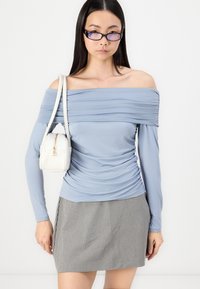 Mėlynas off-the-shoulder viršus su raukiniais, ilgomis rankovėmis, derinamas su pilku languotu mini sijonu. Modelis laiko mažą baltą rankinę.