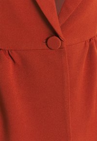 Czerwona bluzka z materiału o dopasowanym kroju, kołnierzem z klapami oraz okrągłym guzikiem w talii, z delikatnymi zakładkami dodającymi tekstury.