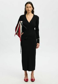 Cardigan nero a coste con scollo a V e bottoni, abbinato a una gonna coordinata. La modella tiene una borsa nera e sciarpe con accenti rossi. Scarpe flat rosse.