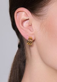 Złote kolczyki w formie obręczy, składające się z trzech splecionych pasków o wypolerowanej powierzchni, mocowane do płatka ucha, podkreślające odcień skóry.