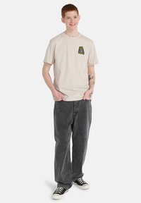 Μπεζ βαμβακερό μπλουζάκι με πράσινο γραφικό στο αριστερό στήθος, συνδυασμένο με χαλαρά σκούρα γκρι τζιν και μαύρα πάνινα παπούτσια.