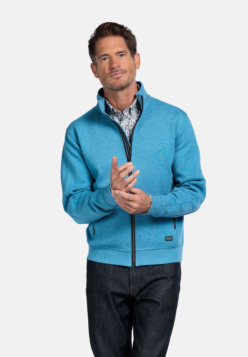 Blauwe zip-up jas van gestructureerde stof, met een hoge kraag, zwarte afwerking en een slim fit, gedragen over een bloemenhemd en jeans.