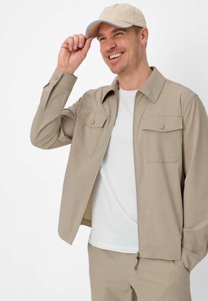 Mand smilende og tipper beige kasket, iført beige jakke med knaplukning på brystlommerne og hvid t-shirt, mod hvid baggrund.