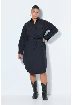 Mujer de cabello corto con vestido tipo camisa negro con cinturón y botas de vaquero negras, de pie contra un fondo blanco con una mano en la cadera.