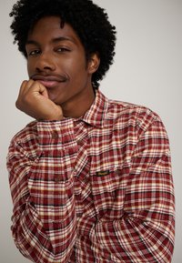 Camicia di flanella a quadri rossi e bianchi con bottoni marroni e una piccola toppa del marchio “Wrangler” sulla tasca sinistra del petto.