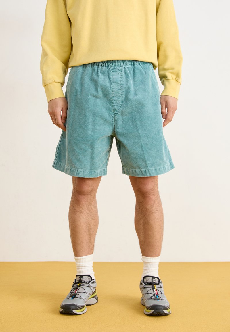 Helle türkisfarbene Cordshorts mit elastischem Bund, die bis zur Mitte des Oberschenkels reichen. Kombiniert mit einem gelben Sweatshirt und grauen Sport Schuhen.