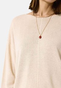 Beige Strickpullover mit weitem Ausschnitt und lockerer Passform. Verfügt über eine mittige Naht und eine schlichte goldene Halskette mit einem rötlichen Steinanhänger.