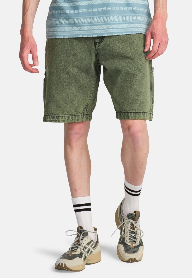 Shorts cargo en jean vert olive avec coutures visibles, poches latérales, aspect usé, associés à des chaussettes blanches rayées et des baskets beige.