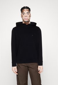 Sweat à capuche noir avec poche kangourou, fabriqué en tissu doux ; présente un petit logo sur la poitrine et des poignets côtelés. Porté avec un pantalon marron.