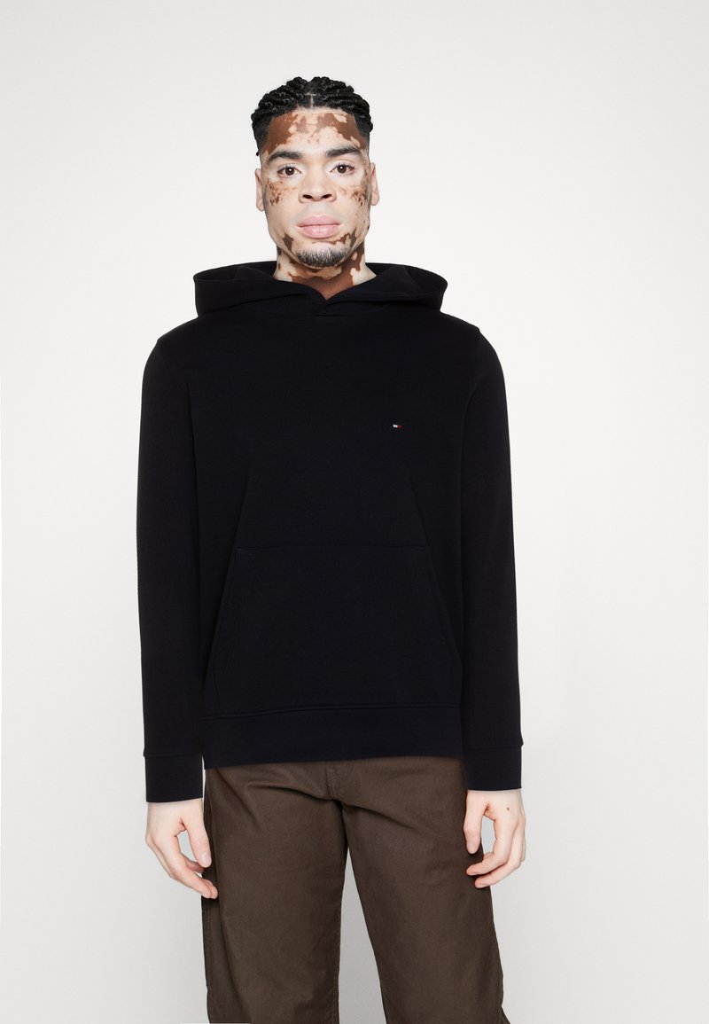 Sweat à capuche noir avec poche kangourou, fabriqué en tissu doux ; présente un petit logo sur la poitrine et des poignets côtelés. Porté avec un pantalon marron.