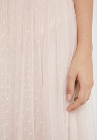 Ljusrosa tyllklänning med ett lagerdesign och vit stjärnmönstrad brodyr. En hand håller tyget nära fållen.