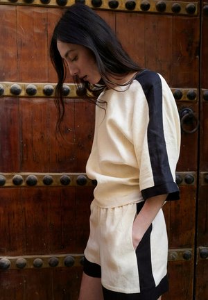 Mujer de cabello largo y oscuro, vestida con un conjunto crema con rayas negras, de perfil con la mano en el bolsillo frente a una puerta de madera.
