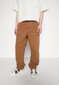 Bruna bomullbyxor med avkopplad passform, med sidofickor, synliga sömmar och lätt avsmalning vid nederkanten, i kombination med vita sneakers.