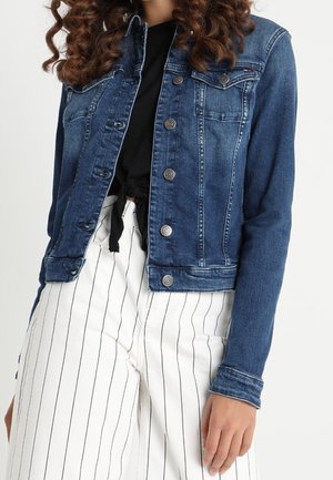 Vrouw draagt een nauwsluitend blauwe spijkerjack over een zwart geknoopt overhemd en witte broek met verticale zwarte strepen.