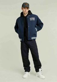 Νεαρός άνδρας με ναυτική ζακέτα, μαύρη κουκούλα, μαύρα παντελόνια, λευκά μποτάκια και σκούρο καπέλο μπέιζμπολ, στέκεται με τα χέρια στις τσέπες.