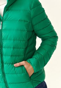 Zelena puffer jakna s kvadratnim šavovima, zatvaranjem na zip, kapuljačom i bočnim džepovima; izrađena od laganog, glatkog materijala.