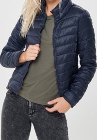 Giacca puffer navy con design trapuntato, colletto alto e zip frontale. Indossata sopra una t-shirt oliva, abbinata a jeans scuri. Texture liscia.